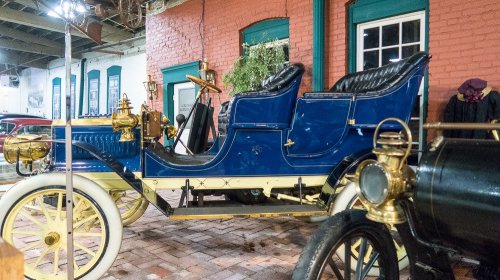 A vintage blue automobile with brass accents, parked indoors against a backdrop of red brick walls.
