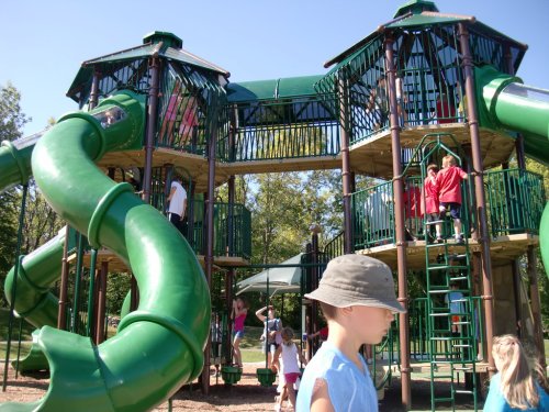 Children play on a large green playground structure with slides and climbing areas under a clear blue sky.