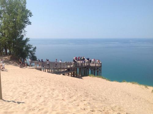 A sandy beach with a wooden pier extending into a calm blue lake, surrounded by trees and people enjoying the view.