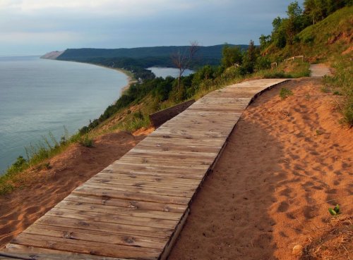 A wooden boardwalk winds along a sandy hillside, overlooking a calm lake and lush green landscape.