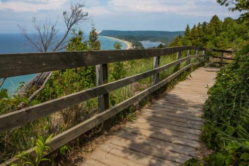 A wooden boardwalk winds through greenery, overlooking a scenic coastline and blue ocean under a partly cloudy sky.