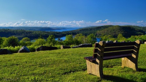 A scenic view of a lake surrounded by green hills, with a wooden bench in the foreground under a clear blue sky.