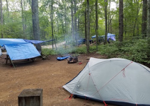 A campsite in a forest with tents, blue tarps, and a small fire pit surrounded by trees. Smoke rises from the fire.