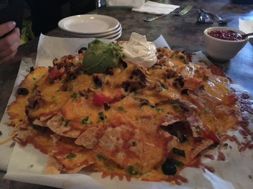A large plate of nachos topped with melted cheese, guacamole, sour cream, tomatoes, and olives on a table.