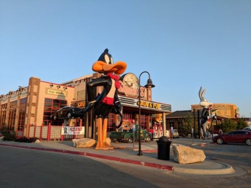 A large Daffy Duck statue stands outside a restaurant, with a vintage car and Bugs Bunny statue nearby.