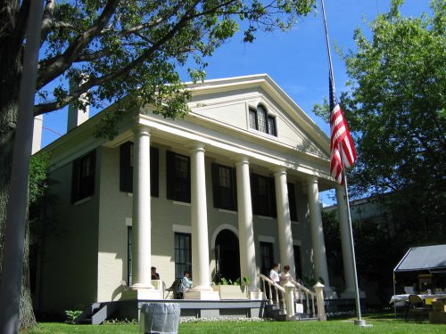 A large, historic house with columns, surrounded by trees and a flagpole, under a clear blue sky.