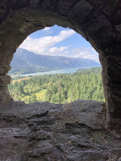 View through a stone archway, showcasing a lush green landscape and mountains under a partly cloudy sky.