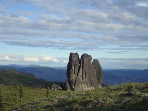 A tall, jagged rock formation stands in a grassy landscape with rolling hills and a cloudy sky in the background.