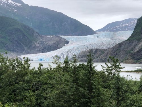 A glacier flows down a mountain, surrounded by lush greenery and a calm lake under a cloudy sky.
