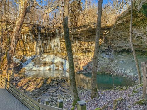 A serene landscape featuring a frozen waterfall, rocky cliffs, and a calm pool, surrounded by trees in winter.
