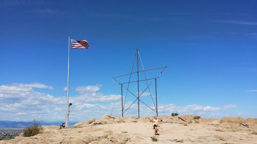 A person stands near a large metal star structure on a rocky hill, with an American flag waving in the background.