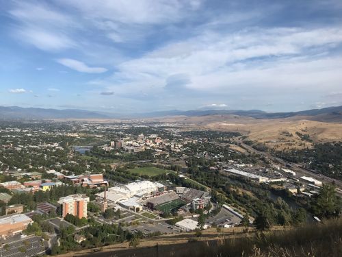 A panoramic view of a city surrounded by hills, with buildings, roads, and a river visible under a blue sky.