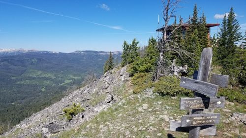 A scenic mountain view with a lookout tower and wooden signposts on a rocky hillside, surrounded by trees.