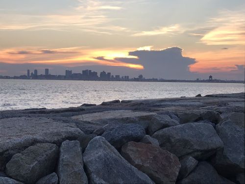 A rocky shoreline with a sunset over a city skyline, featuring clouds and a calm water surface.