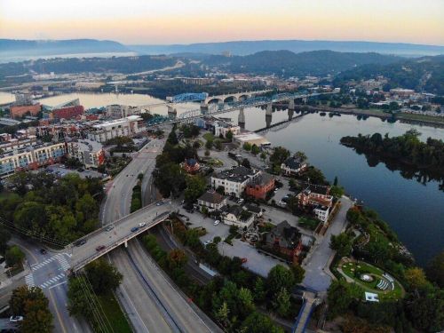 Aerial view of a riverfront city with bridges, buildings, and greenery at sunset.