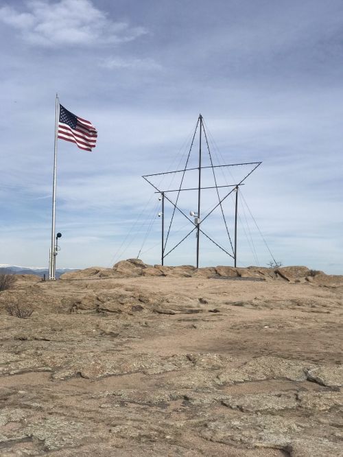 A large metal star structure on rocky terrain, with an American flag waving nearby against a cloudy sky.