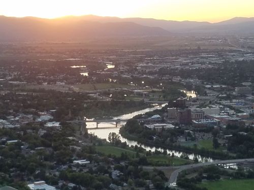 Aerial view of a city at sunset, with rivers, greenery, and mountains in the background.