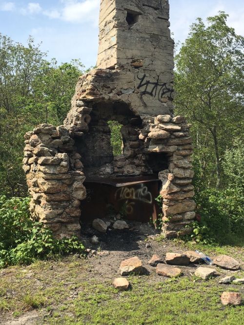 A partially collapsed stone chimney with graffiti, surrounded by greenery and scattered rocks on the ground.