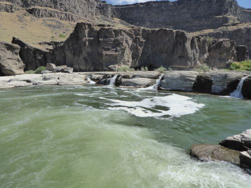 A serene river flows over rocks, surrounded by rugged cliffs and greenery under a clear blue sky.