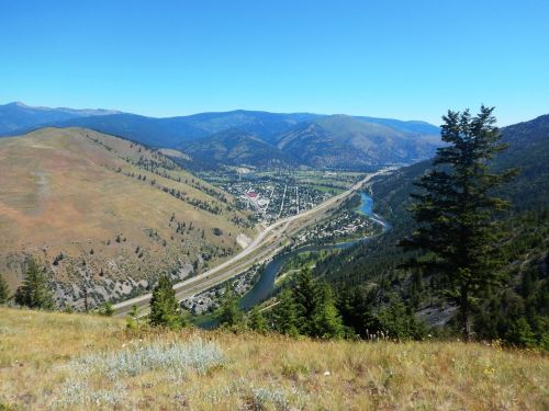 A scenic view of a valley with a river, highway, and mountains under a clear blue sky. Green hills in the foreground.