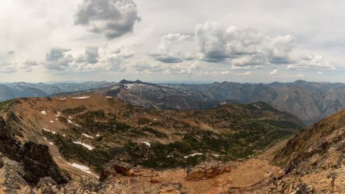 A panoramic view of rugged mountains under a cloudy sky, with patches of snow and green vegetation in the valleys.