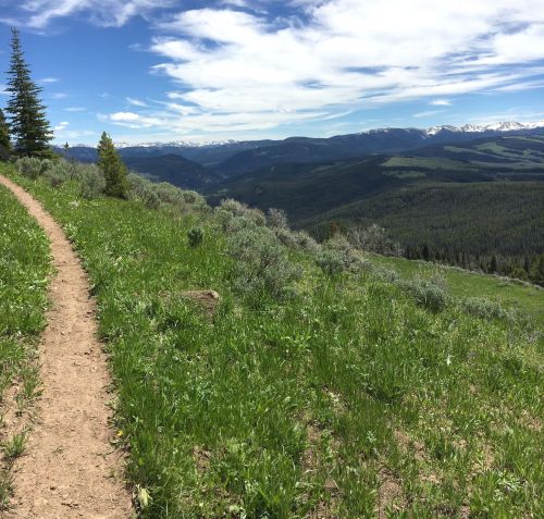 A winding dirt path through lush green grass, overlooking a vast mountainous landscape under a blue sky with clouds.