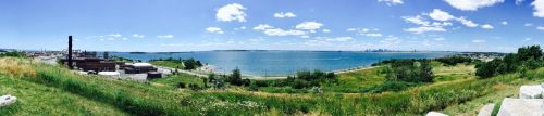 Panoramic view of a lush green landscape overlooking a blue bay and distant city skyline under a clear blue sky.