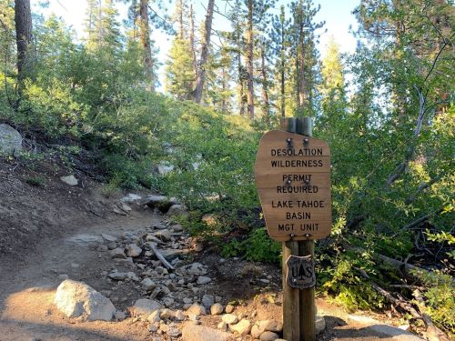 A wooden sign at a trailhead reads "Desolation Wilderness, Permit Required, Lake Tahoe Basin Mgt. Unit," surrounded by trees.