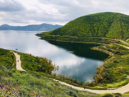 A serene landscape featuring a calm lake surrounded by green hills and a winding path, with a few people walking nearby.