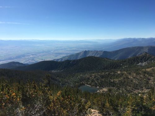 A panoramic view of mountains and valleys under a clear blue sky, with lush greenery and a small lake in the foreground.