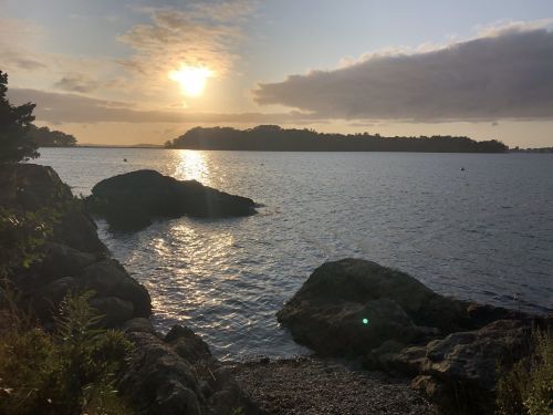 Sunset over a calm water body, with rocky shoreline and silhouettes of distant trees against a cloudy sky.