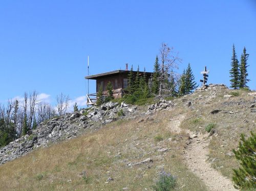 A small wooden cabin on a rocky hill, surrounded by trees under a clear blue sky. A path leads up to the cabin.
