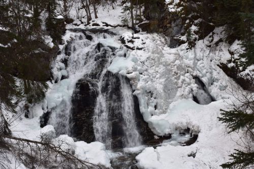 A frozen waterfall surrounded by snow-covered rocks and trees in a winter landscape.