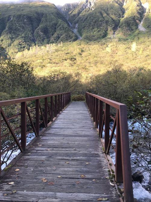A wooden bridge spans a river, surrounded by lush greenery and mountains in the background.