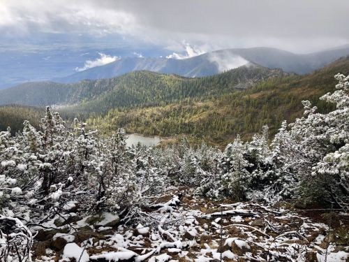 A snowy mountain landscape with evergreen trees, a lake in the valley, and misty clouds overhead.