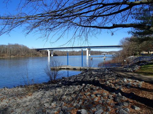 A serene river scene with a bridge in the background, rocky shore, and bare trees under a clear blue sky.