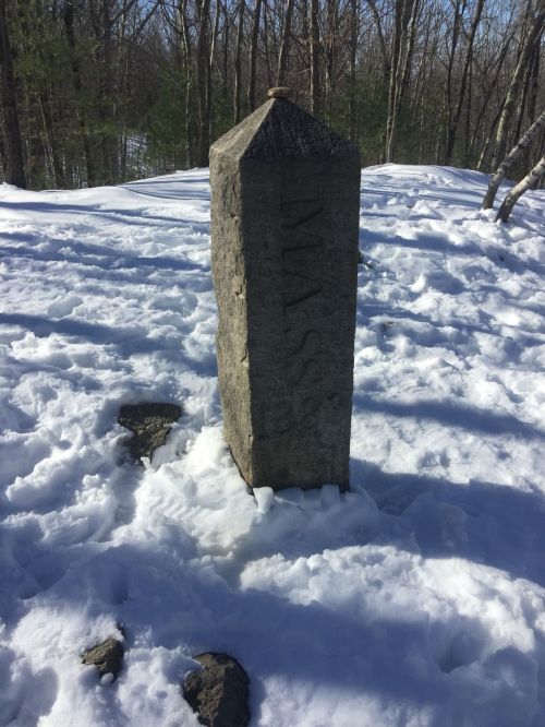 A stone boundary marker labeled "MASS" stands in a snowy landscape surrounded by trees.
