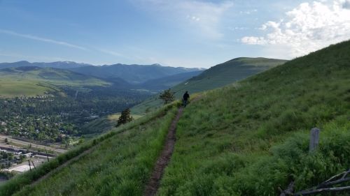 A person walks along a narrow trail on a grassy hillside, with mountains and a valley visible in the background.