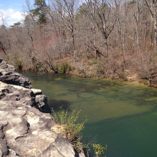 A serene river flows through a wooded area, with rocky banks and bare trees reflecting a peaceful natural landscape.