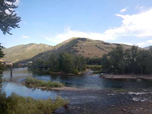 A serene river flows through a valley, surrounded by green hills and trees under a blue sky with scattered clouds.