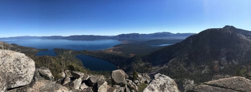 A panoramic view of a serene lake surrounded by mountains and forests under a clear blue sky.
