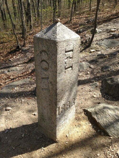 Granite boundary marker with the inscription "P.J." and the year "1833," surrounded by a wooded area.