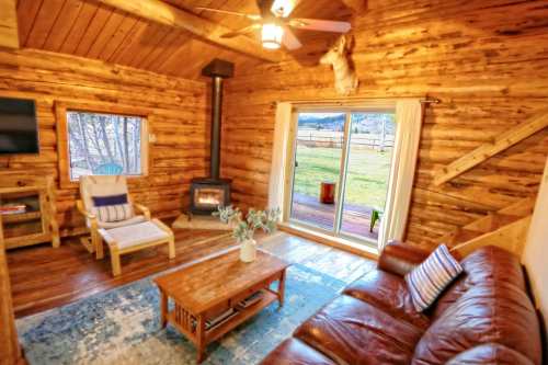 Cozy log cabin interior with a leather sofa, wooden furniture, and a fireplace, overlooking a scenic outdoor view.