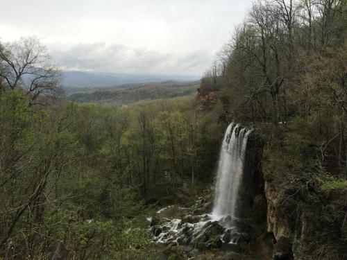 A serene waterfall cascades down rocky cliffs, surrounded by lush green trees and a misty mountain backdrop.