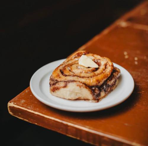 A freshly baked cinnamon roll on a white plate, topped with a pat of butter, resting on a wooden table.