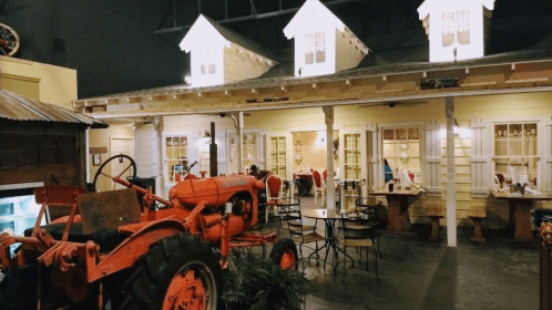 A rustic dining area featuring a vintage orange tractor, wooden tables, and a charming house facade.