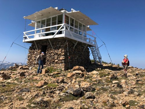 A stone fire lookout tower on a mountain, with two hikers nearby against a clear blue sky.