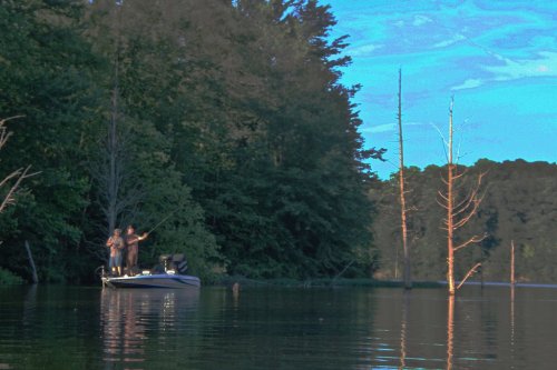 Two people fishing from a boat on a calm lake, surrounded by trees and a clear blue sky.