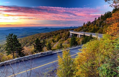 Scenic view of a winding road through colorful autumn trees, with a vibrant sunset over distant mountains.