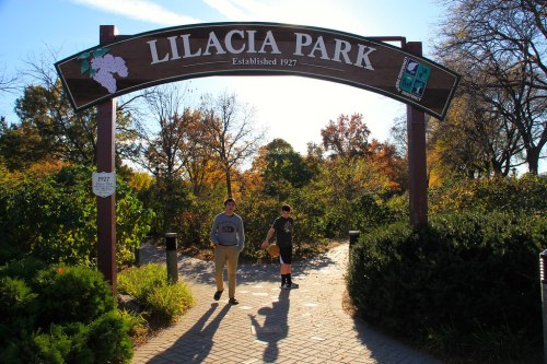 Two people walk under the entrance sign of Lilacia Park, surrounded by trees and autumn foliage. Established 1927.